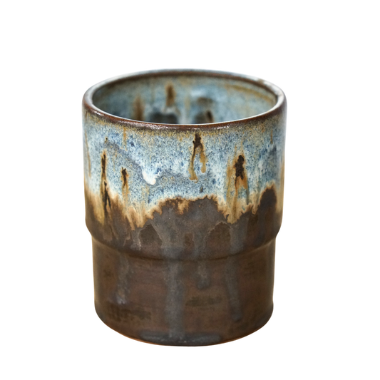 A handmade ceramic cup with a textured, ribbed exterior and a dripping glaze showing shades of brown, blue, and beige on a white background.